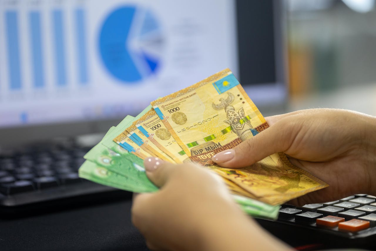 About Human hands counting Kazakh tenge bills near a laptop displaying financial graphs.